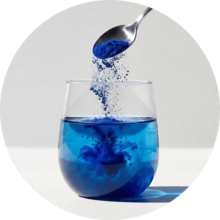 A spoon of blue spirulina powder being tipped into a glass of liquid, dying the liquid a vivid blue